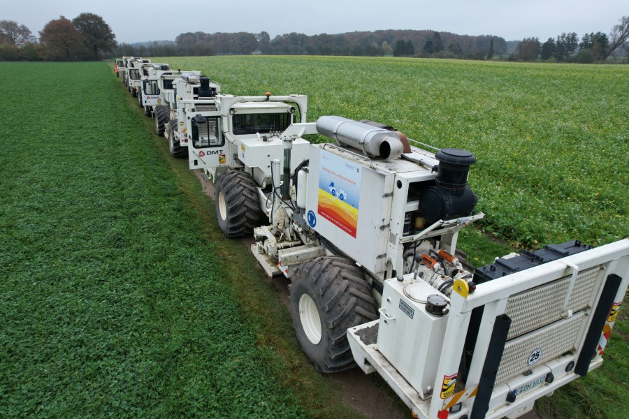 Mehrere weiße Seismik Vibro-Trucks in Reihe auf grünem Feld für geophysikalische Bodenuntersuchungen der Stadtwerke Kaiserslautern.